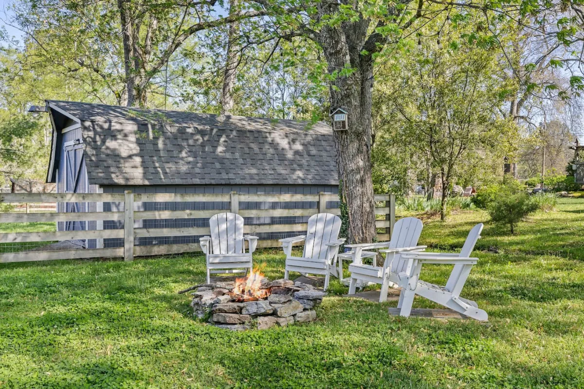 An inviting outdoor fire pit area featuring a circle of Adirondack chairs around a crackling fire, set beneath a large tree with a rustic shed visible in the background. The golden light suggests a tranquil evening atmosphere, perfect for relaxing and enjoying the countryside.