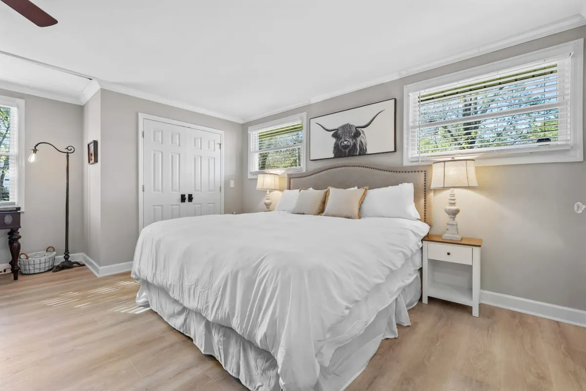 This inviting bedroom features a king-size bed with soft white bedding, framed by large windows that let in abundant natural light. Above the bed, striking bull artwork adds a rustic touch, while table lamps on either side create a softly lit, intimate ambiance. The overall mood is serene, with peaceful garden views visible through the windows.