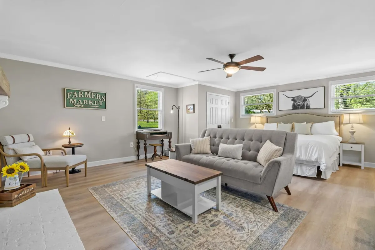 A cozy open-concept space featuring a sofa, coffee table, and a bed, all bathed in gentle morning light. The area is decorated with a farmer's market sign and bull artwork, providing a harmonious blend of rustic and homey elements. Large windows reinforce the airy, light-filled atmosphere.