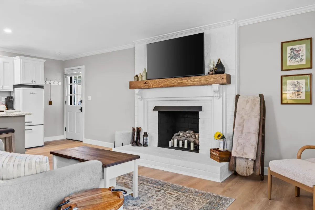 A welcoming living area with a fireplace as the centerpiece, a TV mounted above a rustic wooden mantel, and a cozy coffee table in front. Soft, warm lighting and decorative artwork contribute to the inviting atmosphere, setting the scene for relaxing evenings or intimate gatherings.