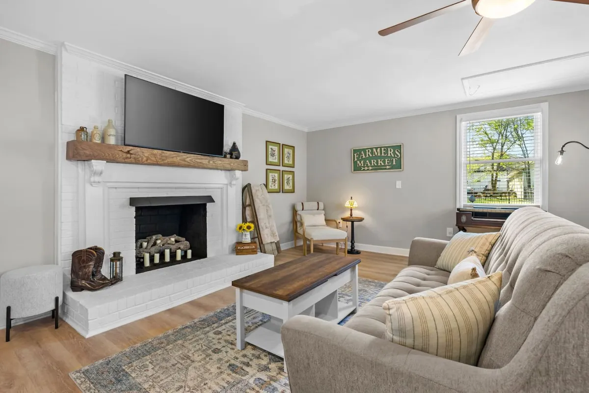 This inviting studio interior features a glowing candlelit fireplace as the centerpiece, with a plush sofa and coffee table arranged for comfort. Sunlight streams through a window, offering views of the surrounding gardens and countryside, creating a cozy and tranquil atmosphere perfect for relaxing or connecting with loved ones.