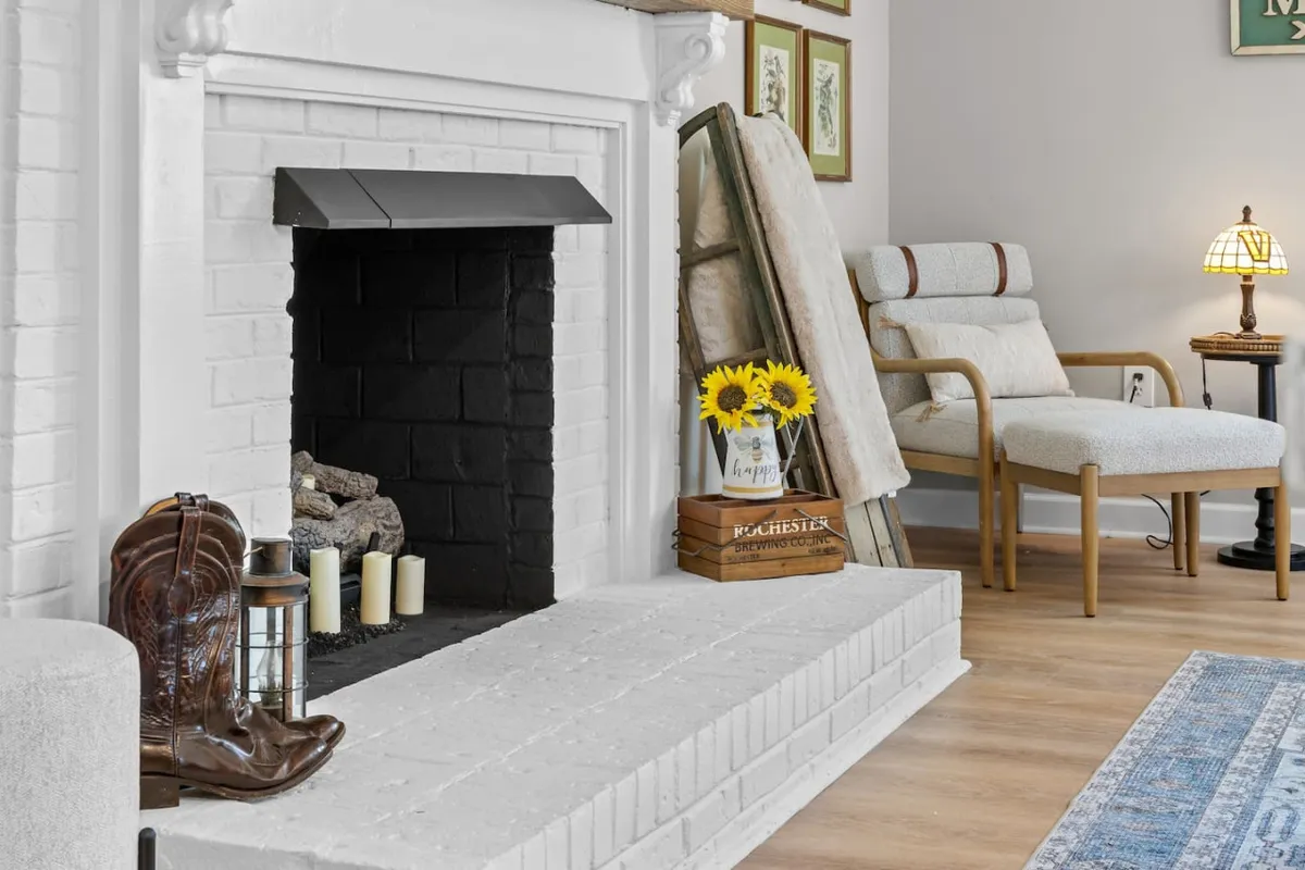 A warmly lit interior featuring a classic fireplace flanked by sunflowers, an upholstered armchair, a lantern, and a pair of boots. The space radiates a sense of comfort and intimacy, creating an ideal setting for quiet relaxation or romantic conversation.