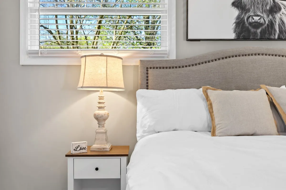 A serene bedroom with a bed dressed in white linens, a glowing table lamp, a decorative sign reading 'Love,' and peaceful artwork that evokes calm. Natural light filters in through the window, creating a tranquil atmosphere.