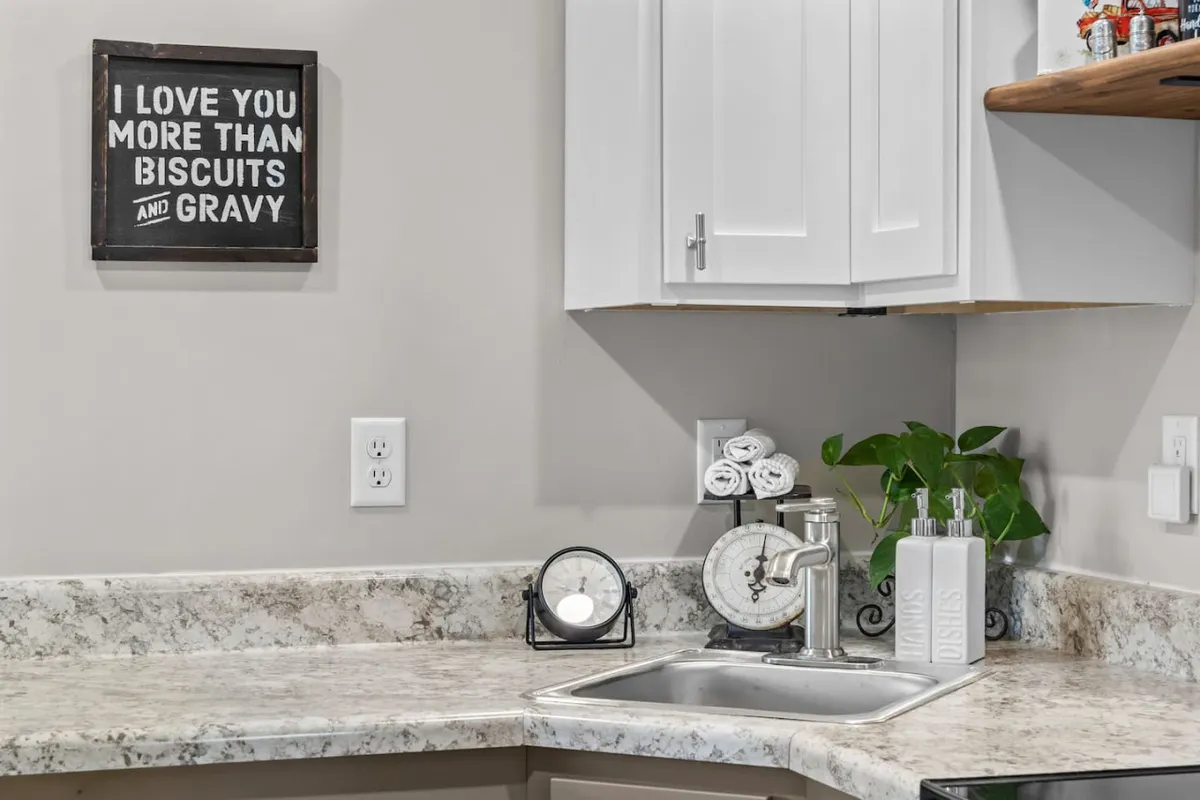 A warm, homely sink area with a decorative clock, framed sign, potted plant, and soft lighting. This functional space is inviting for breakfast conversations and cozy dinner preparations.