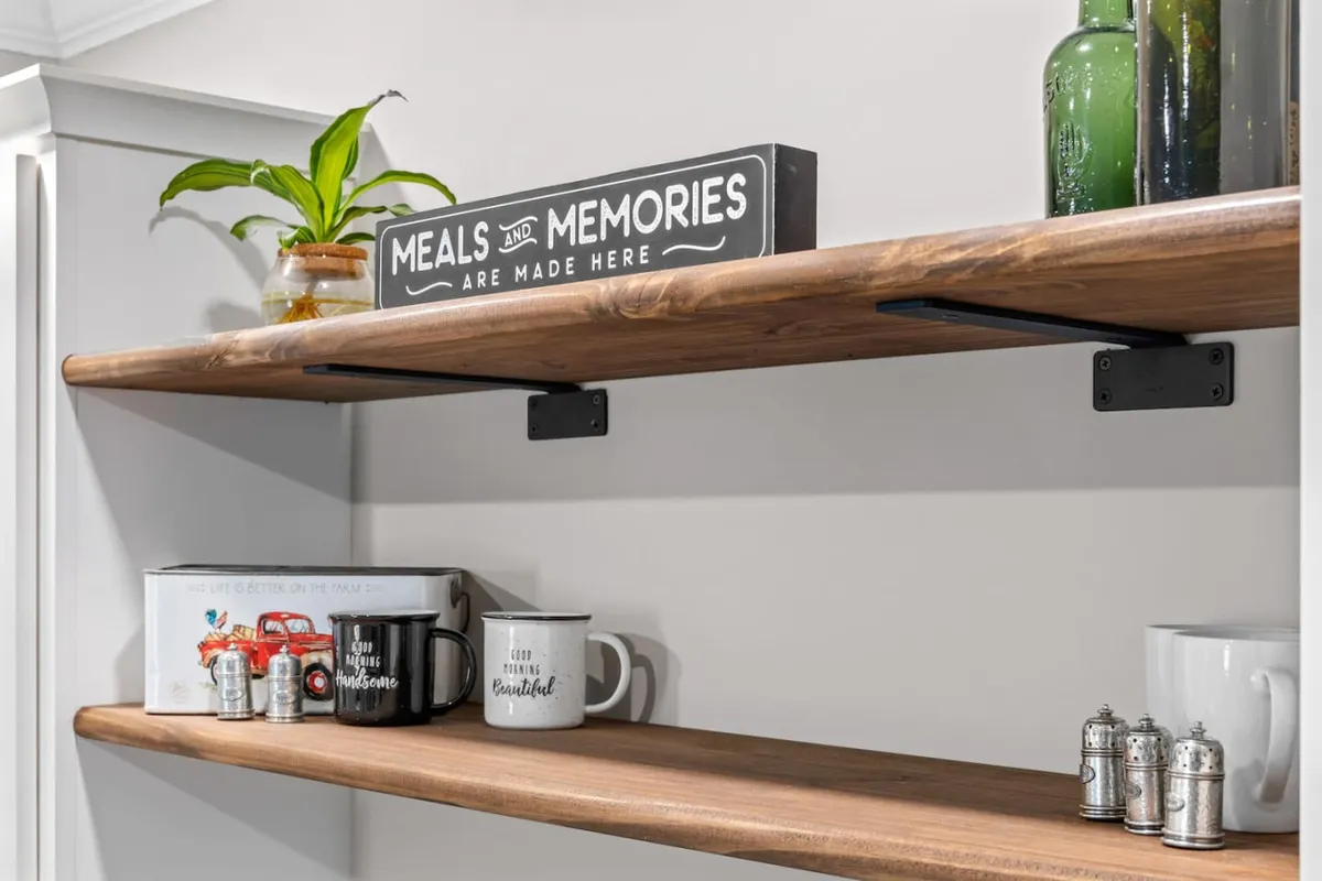Wooden shelves adorned with mugs, glass bottles, a decorative plant, and a charming sign reading 'Meals and Memories are made here.' The warm, nostalgic styling evokes feelings of togetherness and comfort.