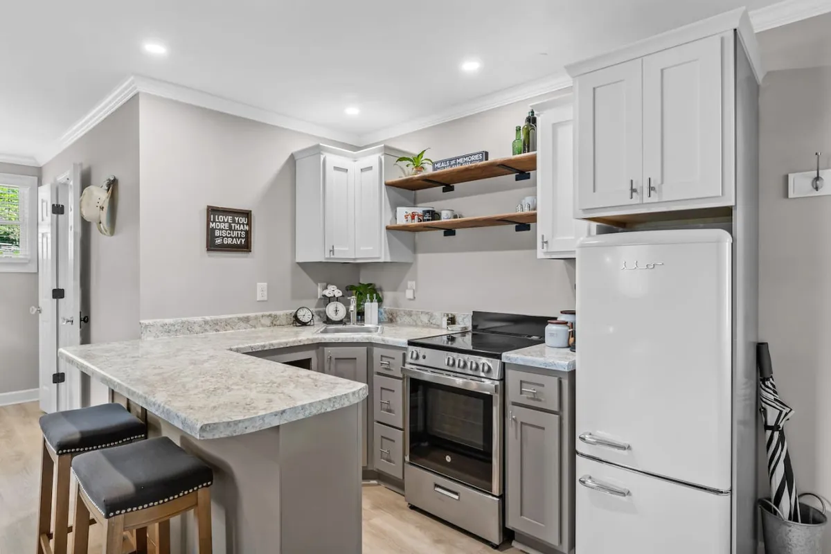 A stylish kitchenette with a kitchen island and bar stools, open shelves adorned with plants, and sleek stainless steel appliances. Granite countertops and thoughtful decor make the space both functional and visually appealing, perfect for preparing meals or sharing coffee.