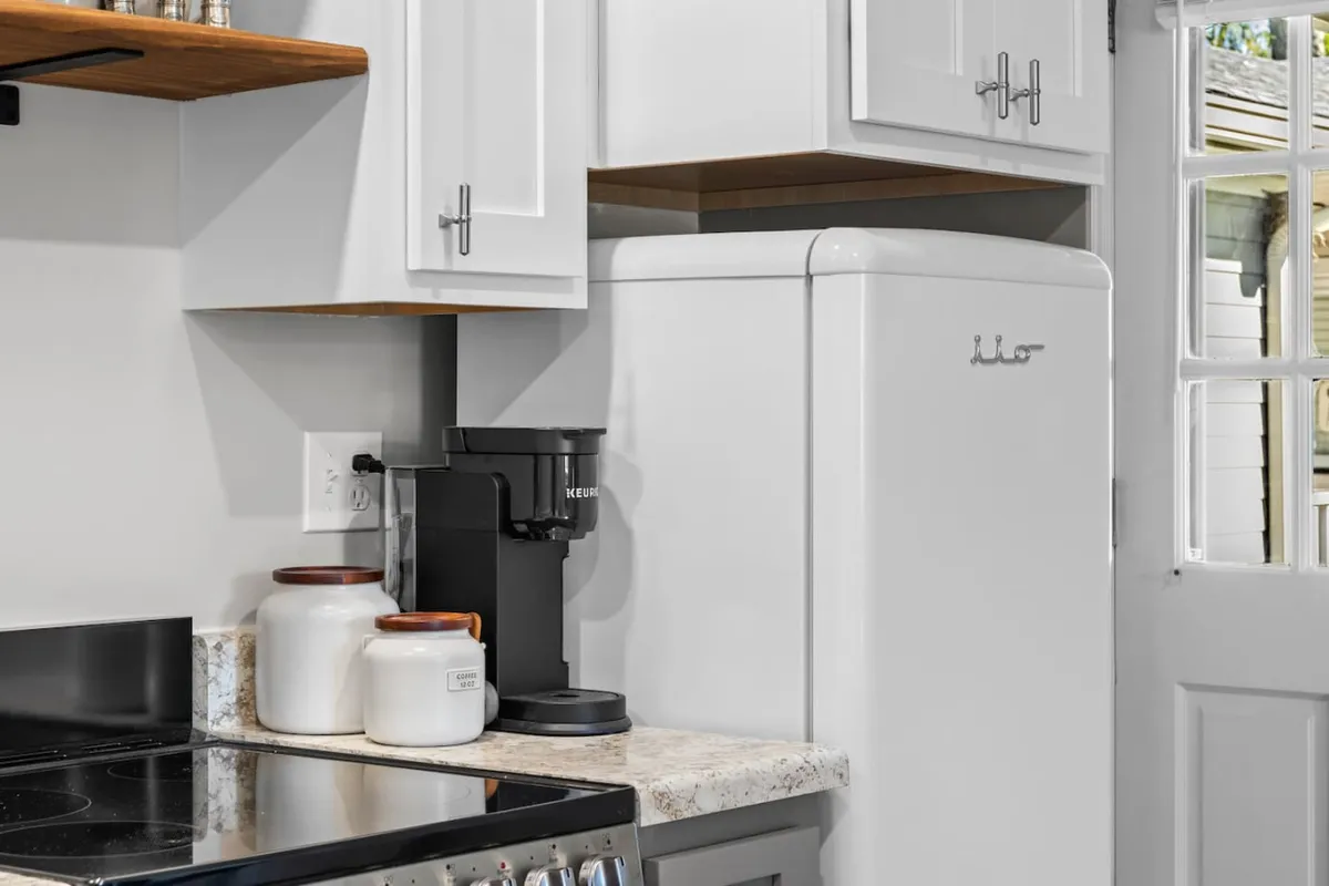 A cozy and modern kitchenette with a stove, coffee maker, white refrigerator, and glass storage jars. The space is bathed in soft lighting, highlighting the clean countertops and practical layout ideal for meal prep and morning routines.